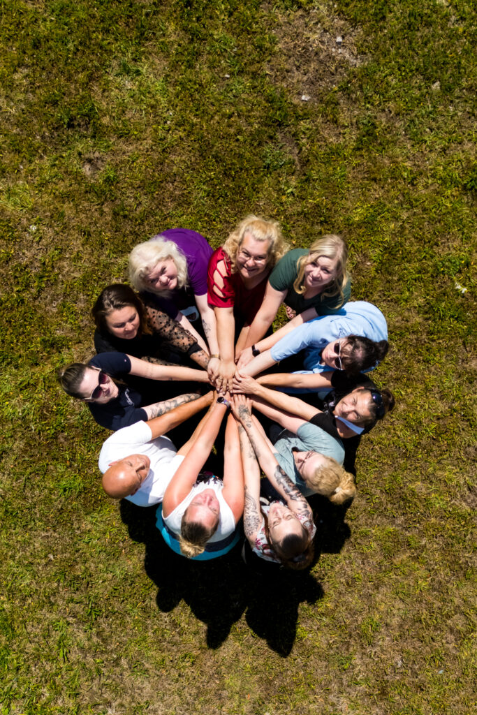 Team von Janine's Pflegeteam liegt im Kreis auf der Wiese und legt die Haende zusammen